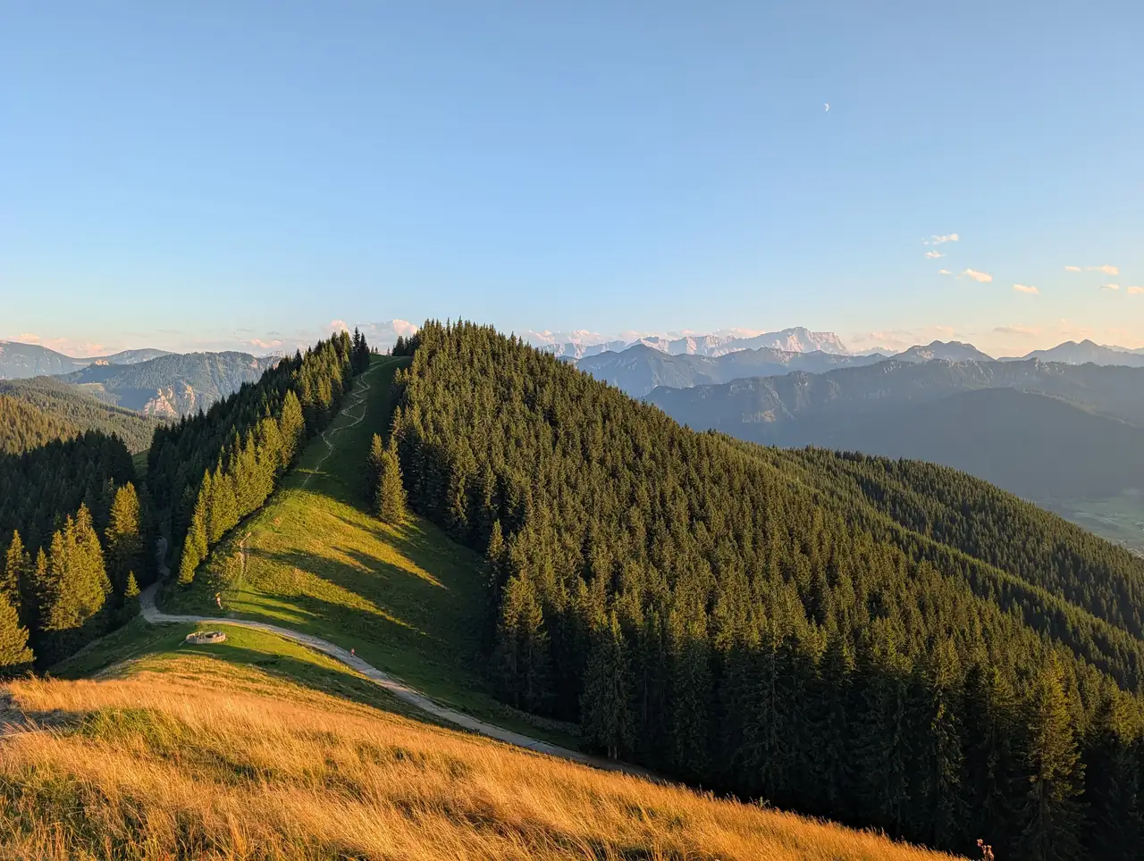 Blick vom Hörnle-Grat bei Bad Kohlgrub über bewaldete Bergrücken und einen Wanderweg bis zur Zugspitze im Abendlicht.